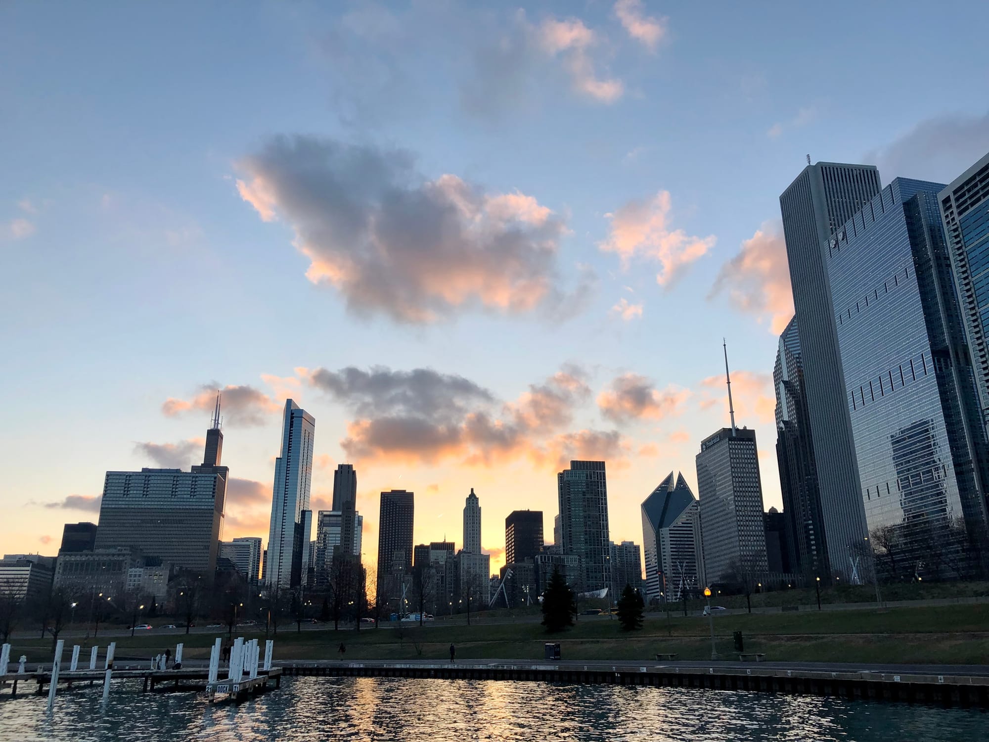 Clouds illuminated in shades of orange, purple and blue over tall buildings and a pond. 