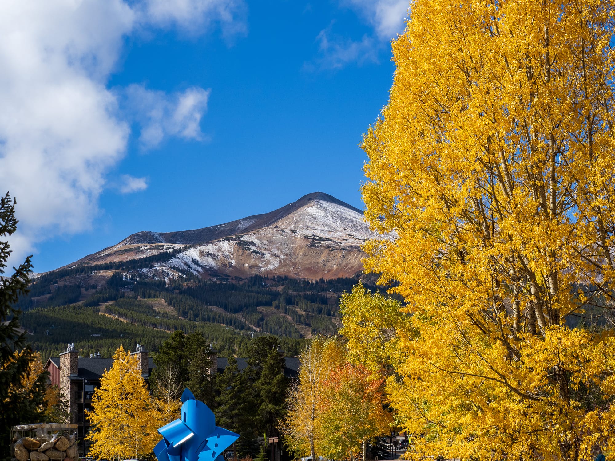 A mountain view with bright yellow fall leaves on one side, blue sky with clouds on the left, and a snow dusted peak in the center. A bright blue sculpture is visible in the foreground.