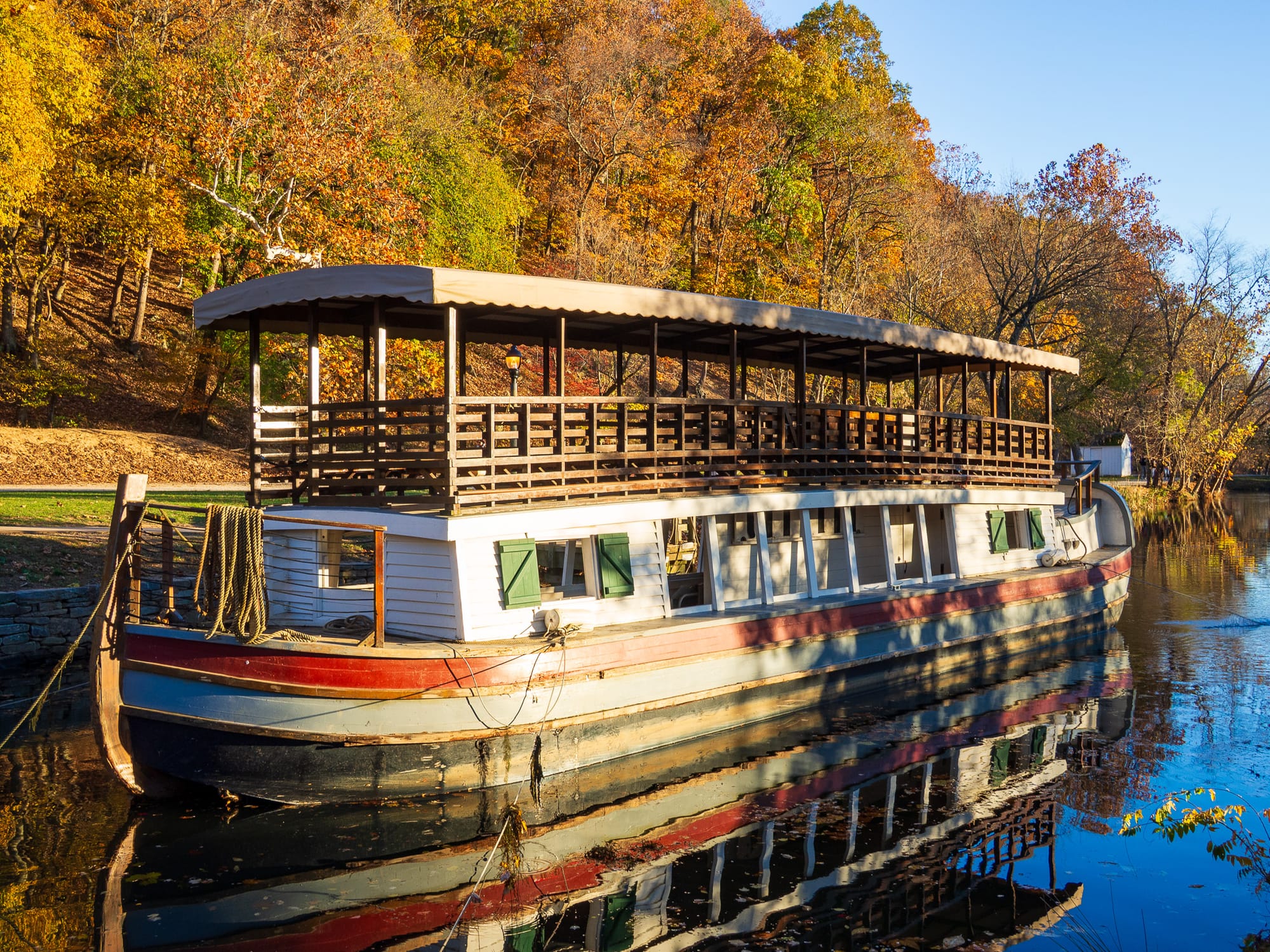 An eighteenth century canal boat sits in reflective waters, with golden hour lit autumn colored trees above.