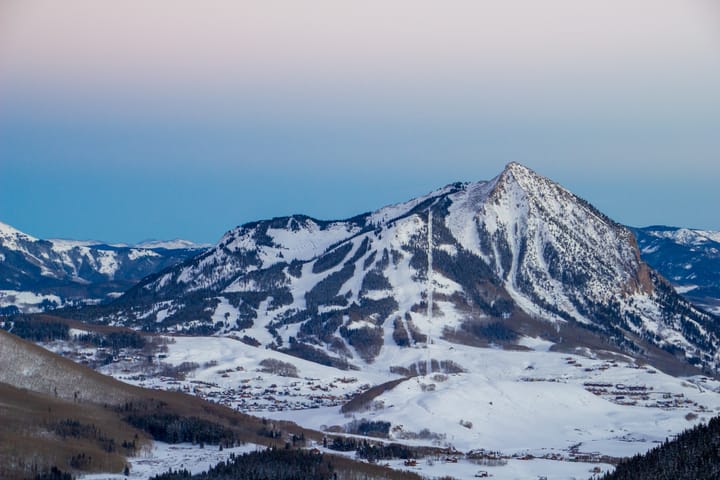 A snowy, craggy mountain cut with ski trails is illuminated in shades of purple as the sun sets. 
