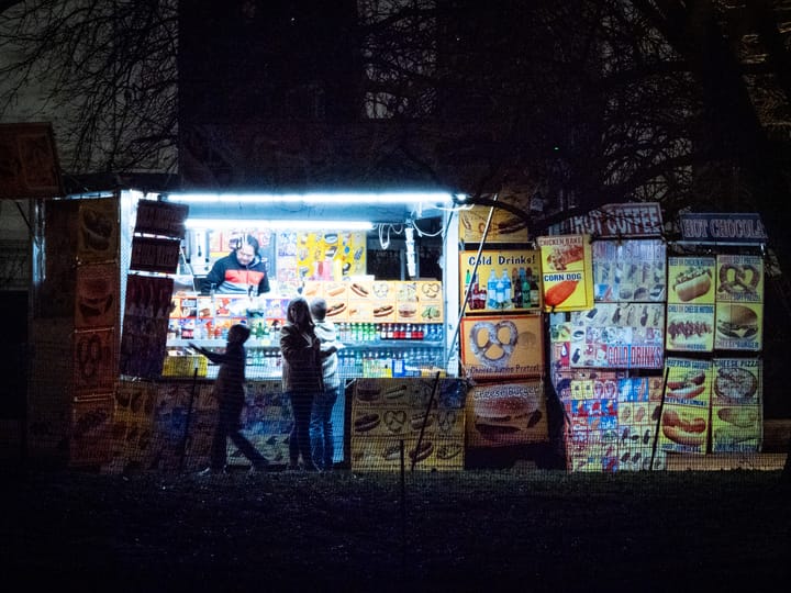 In a night scene, a food truck operator is illuminated by cool blue florescent lights while a family makes their selections. Signs on the right offer cold drinks, pretzels, and hot dogs. 