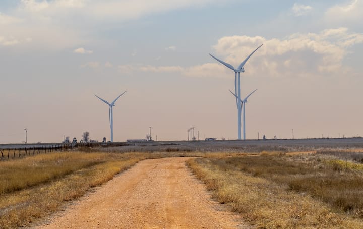 A dirt road in the foreground leads toward three large wind turbines under a hazy sky. 
