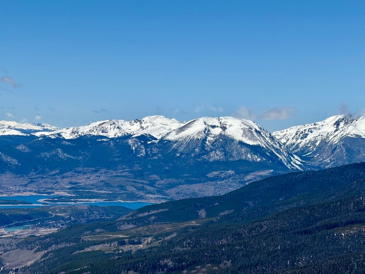 Under blue skies, a tree covered mountain slopes into a mountain valley with a lake visible. Beyond the lake, snow capped peaks rise. 